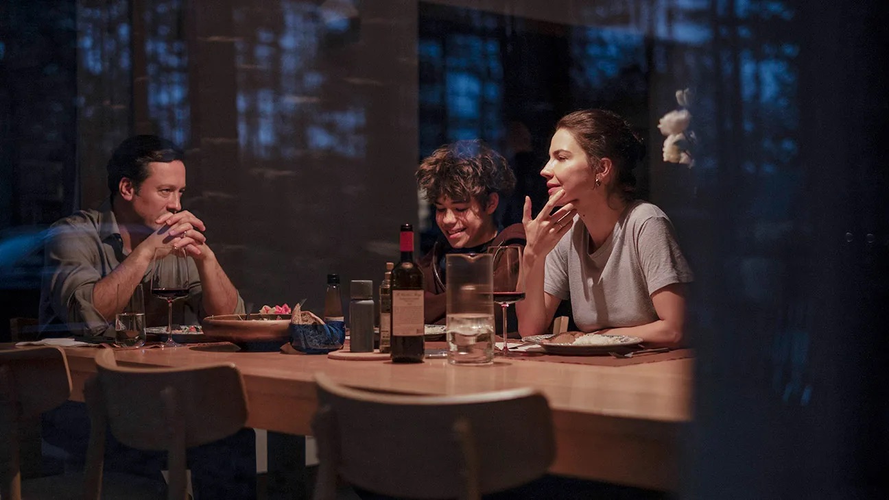 Gleb, Galina e o filho sentados à mesa durante um jantar, vistos através de uma janela que reflete as árvores do lado de fora.