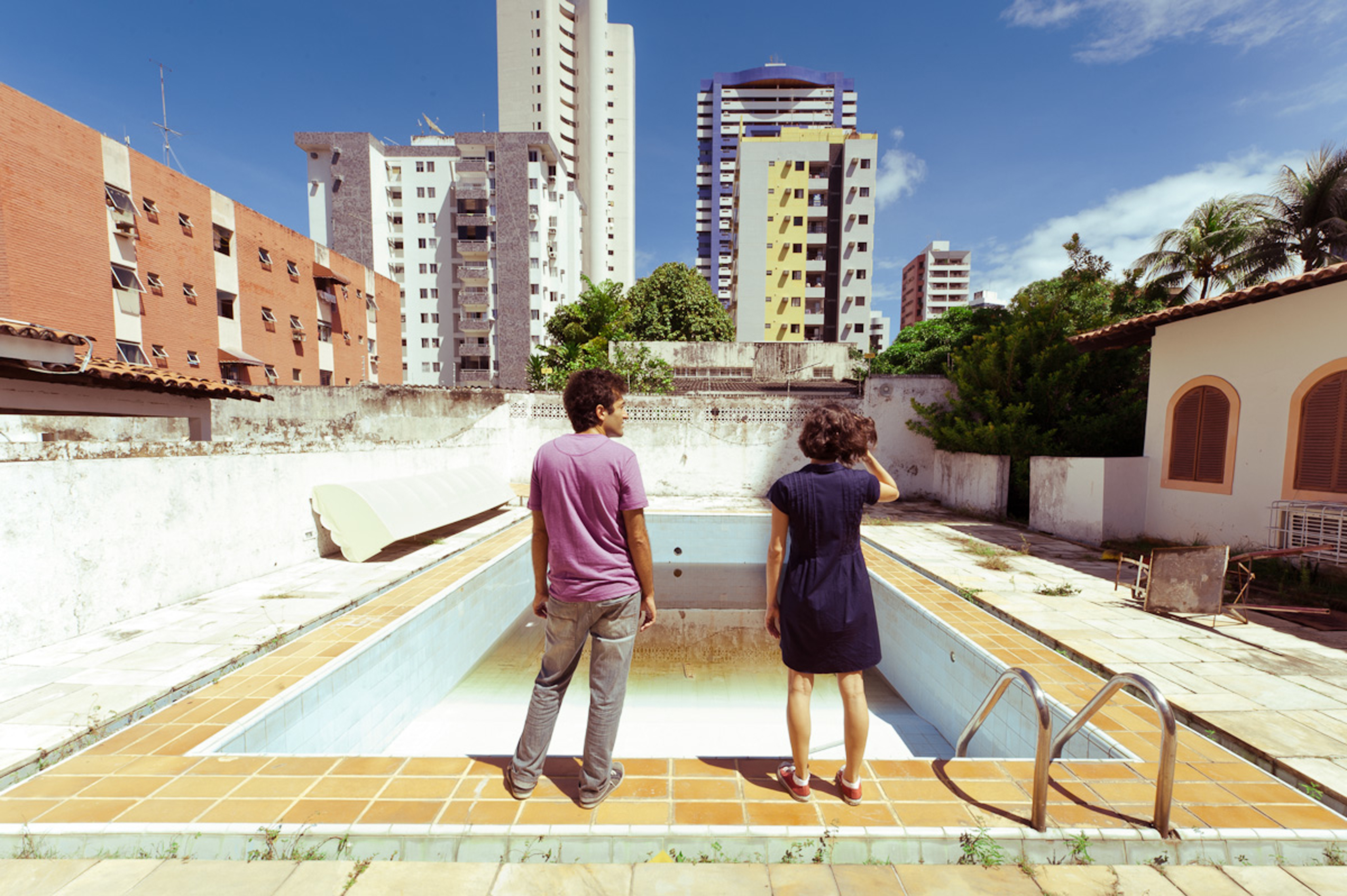 João e Sofia observam uma piscina vazia, com antigos imóveis e edifícios altos ao fundo, em O Som ao Redor.